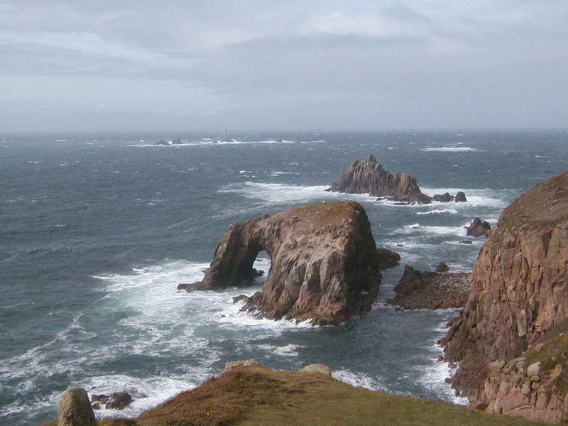 Huge rock arch just off the coast, in the sunshine, rough sea with lots of white spray.