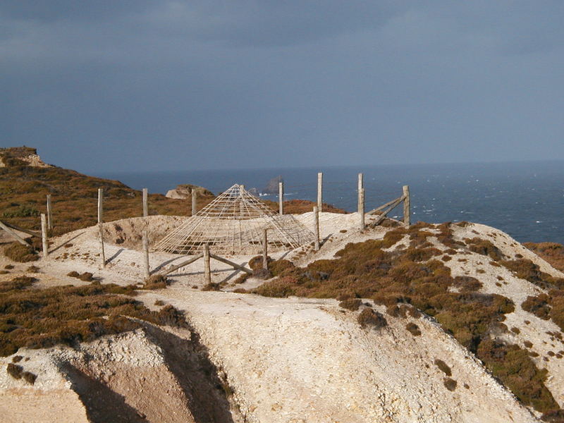 Sandy area with conical metal mesh covering a large hole, sea in the background.