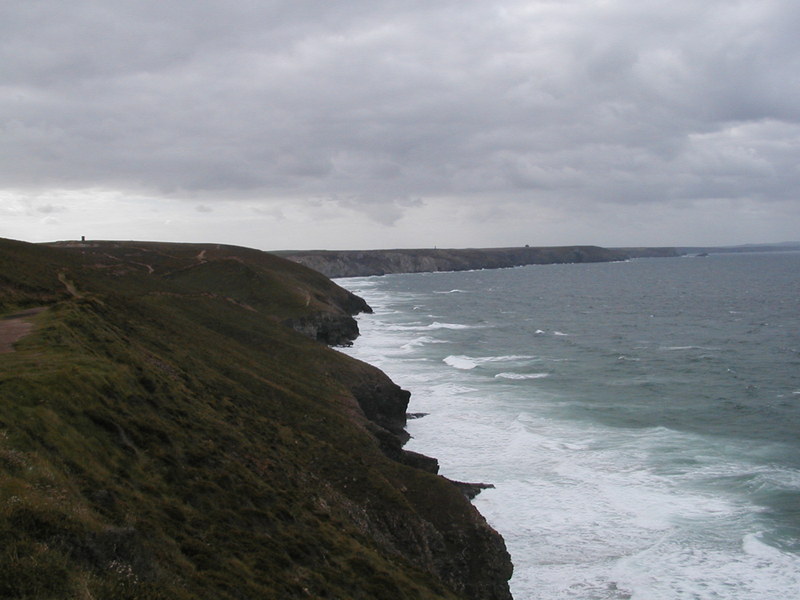 Grey clouds over grassy hillside falling away to small sea cliffs, choppy sea with lots of white surf.