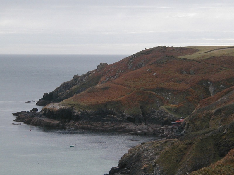 Slipway with front of small orange lifeboat poking out, in front of fern covered hillside descending to rocky shoreline