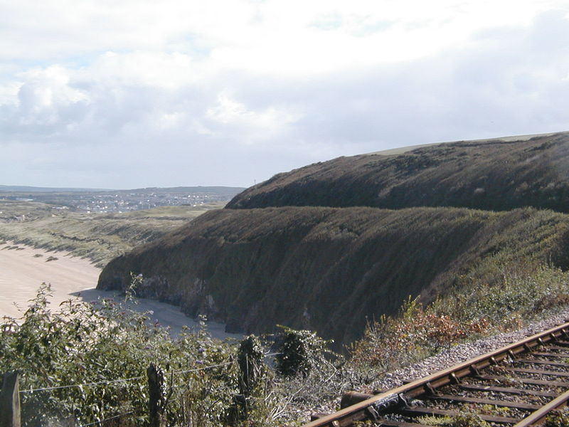 View over railway line of steep, almost vertical, tree covered hillside running down to the beach. Small path just visible traversing right on the edge!