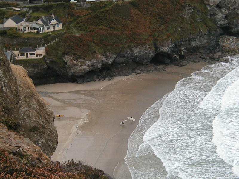 View down to a beach from cliffs, two small surfers far below about to walk into the sea