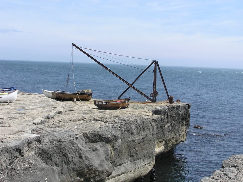 Rusting hand winched crane and a handful of small boats on top of a limestone cliff.