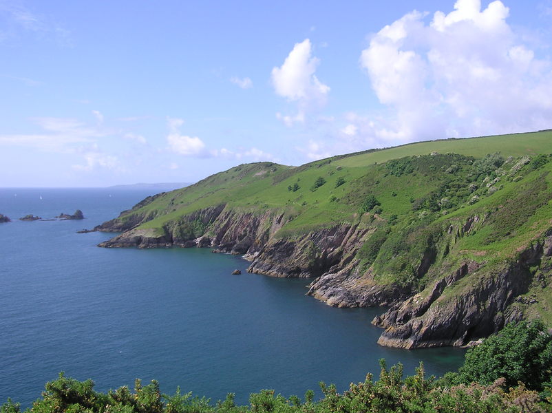 Looking out towards headland, lots of rocky inlets at the shoreline, grassy hillside above, blue sky with a few fluffy white clouds.