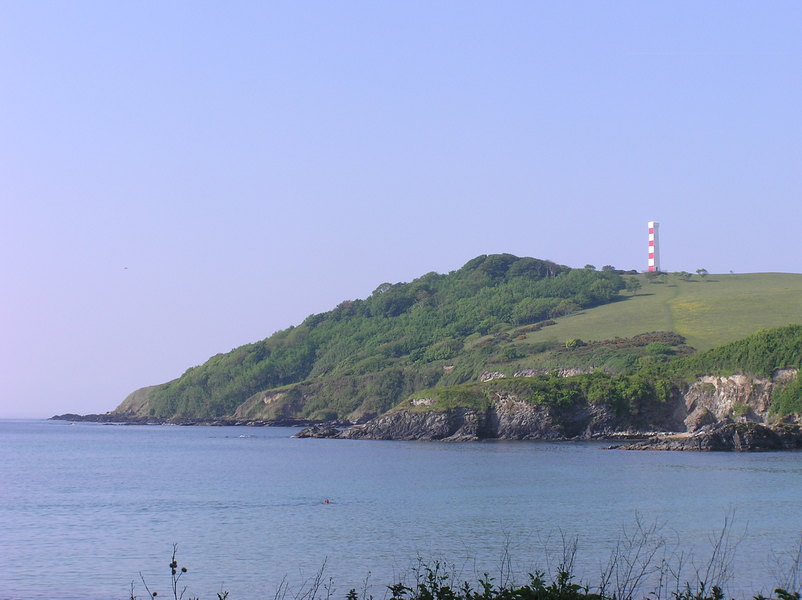 Looking back to headland, covered in trees gently sloping down to the sea, red and white striped marker on top, smooth flat sea and blue sky.