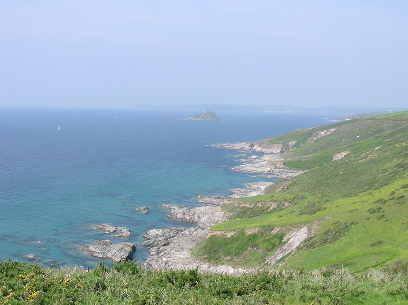 Gently sloping grassy hills leading down to brown rocky shoreline, calm sea, and blue hazy skies in the distance.