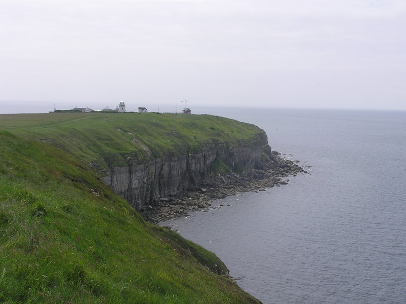 Vertical limestone cliffs rising up from the sea, grassy top and small lighthouse sticking out.